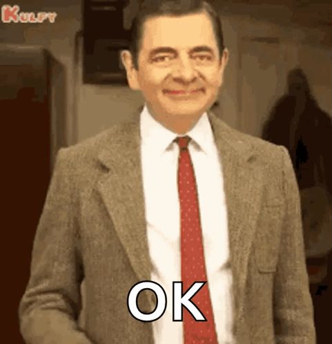 mr bean is wearing a suit and tie and smiling while saying ok .