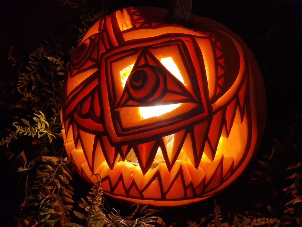 Ornately carved pumpkin. One eye is a square with a triangle iris then round pupil in the middle. The other is a triangle with a square iris and round pupil. The brow line is alternating triangles. It has sharp teeth in a big wide grin, each of the individual teeth have also have triangles carved out, leaving only an outline.
