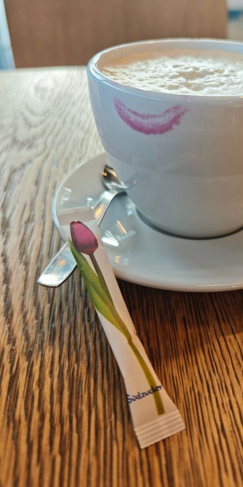 Eine große weiße Tasse mit Milchkaffee und einem pinkfarbenen Lippenstiftabdruck. Auf der Untertasse liegt neben dem kaffeelöffel ein Zuckertütchen, auf dem eine rosafarbene Tulpe abgedruckt ist.