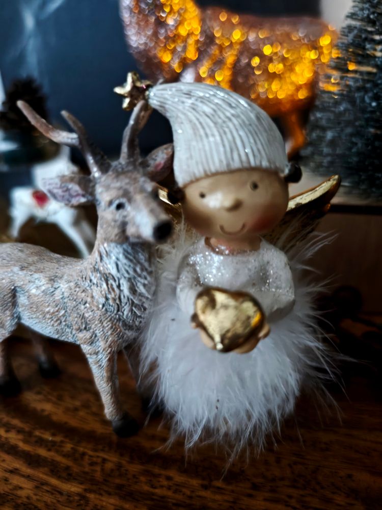 Deko-Hirsch Rudolph steht neben einem kleinen Engelchen, das ein kleines goldenes Herz in die Kamera hält. Im Hintergrund sieht man die Umrisse eines weißen Schaukelpferds und eines goldschimmernden großen Hirsches.