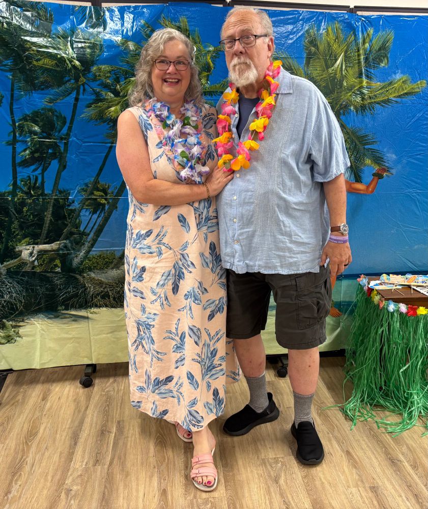 A senior couple dressed for a luau. The woman is wearing a sherbet colored maxi-dress and a blue lei. The man is wearing shorts and an island shirt with a pink and yellow lei. 