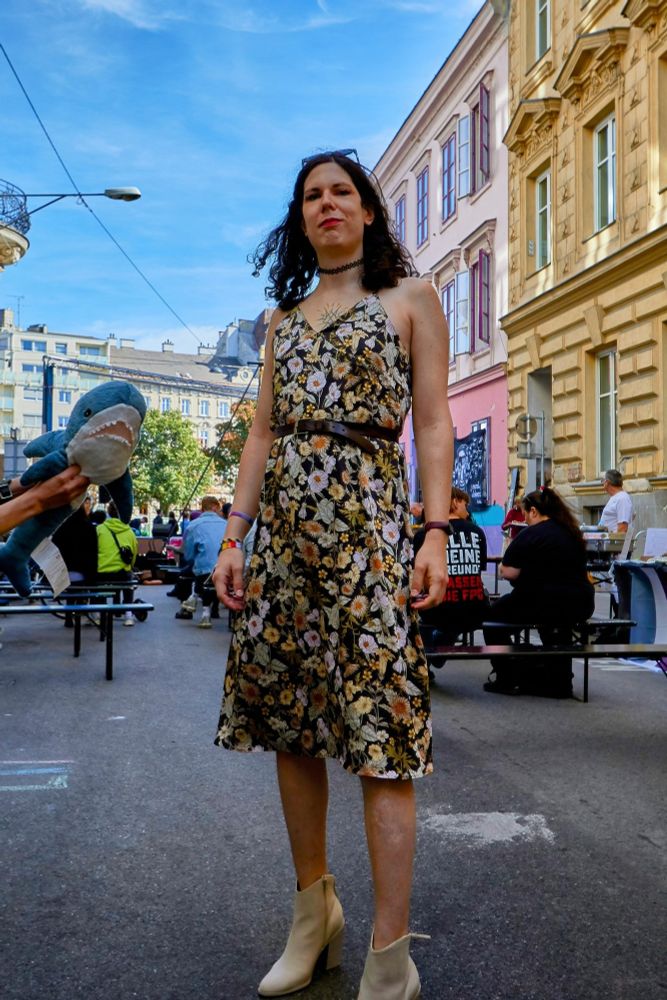miré being hot af, wearing a knee-length sleeveless floral dress with heeled light grey ankle boots. a person is holding a small blåhaj on the left side of the pic.