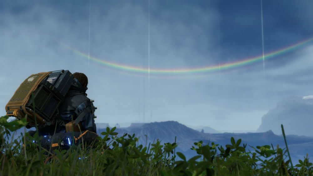 a screenshot of a Death Stranding. Sam is posted up on a rock side in the grass to take a gander at the rainbow.