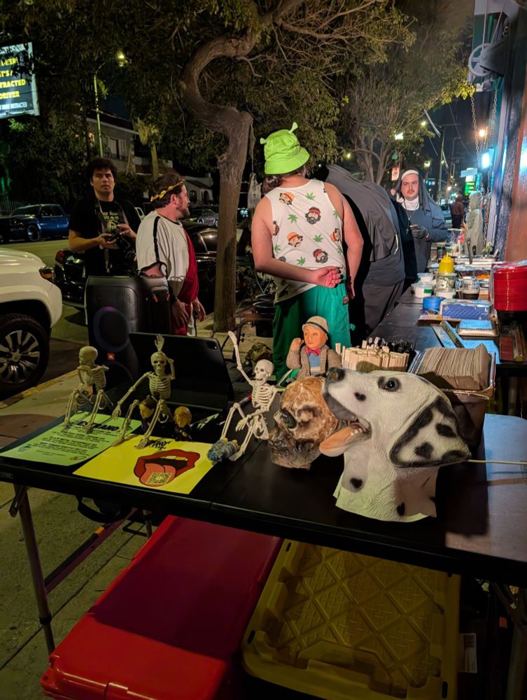 The front of the line for the Estrano Things pop up. The service counter is littered with miniature skeletons, a skull, a dog mask, and other odds and ends. The man himself, Diego Argotti, stands with his back to the camera with a Cheech and Chong tank top and Shrek hat (Shrek themed crocs not pictured). Around him are other chefs and cooks, most of whom are in costume as nuns and one stands in a yoga. A professional photographer stands in the background facing the camera.
