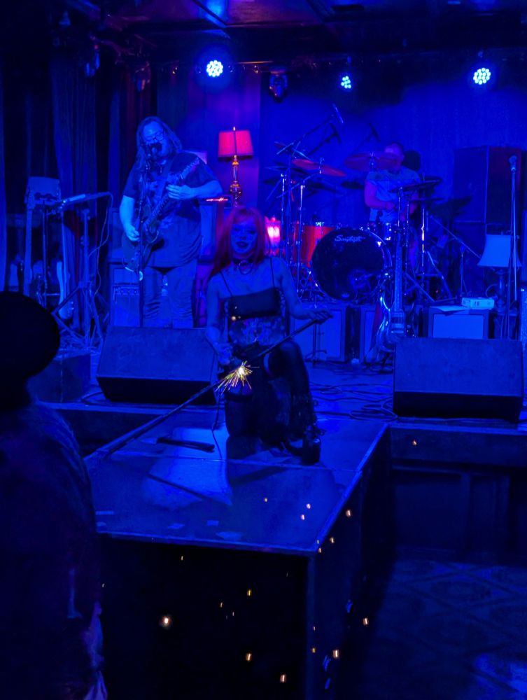 Violet Noirr, using an angle grinder to shoot sparks off a metal poll, while the band The Pretty Pils plays behind her.