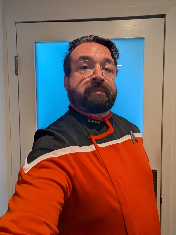 Me, in a Lower Decks command red jacket standing in front of a white door with a large window taking up its center.
