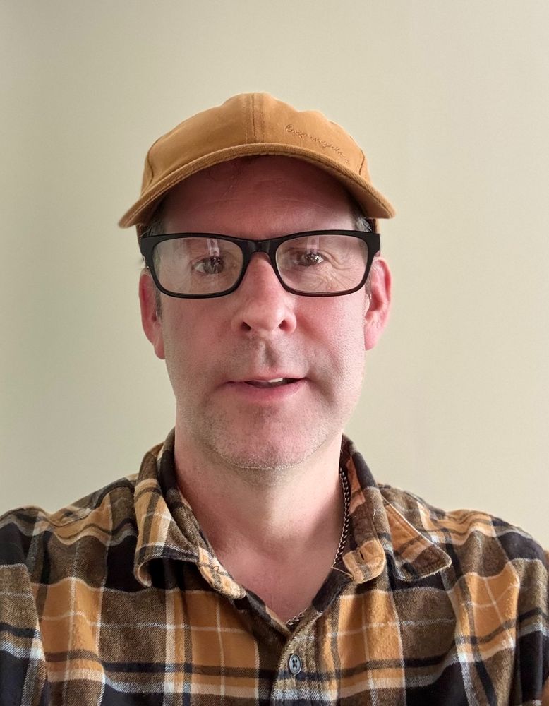 A photo of Lloydi facing the camera, wearing a tan baseball cap, dark-rimmed glasses, and a plaid shirt, looking thoughtful and ready for some in-depth accessibility conversations