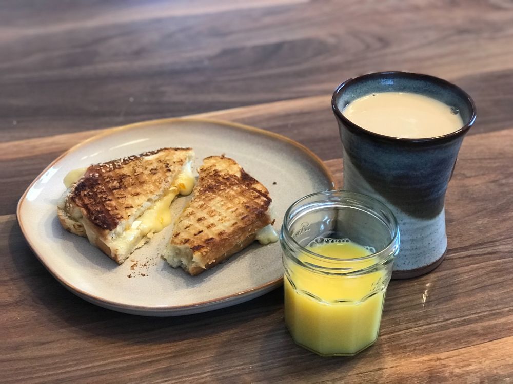 An apricot and brie grilled cheese on a plate beside a repurposed jam jar filled with orange juice and a large mug of tea