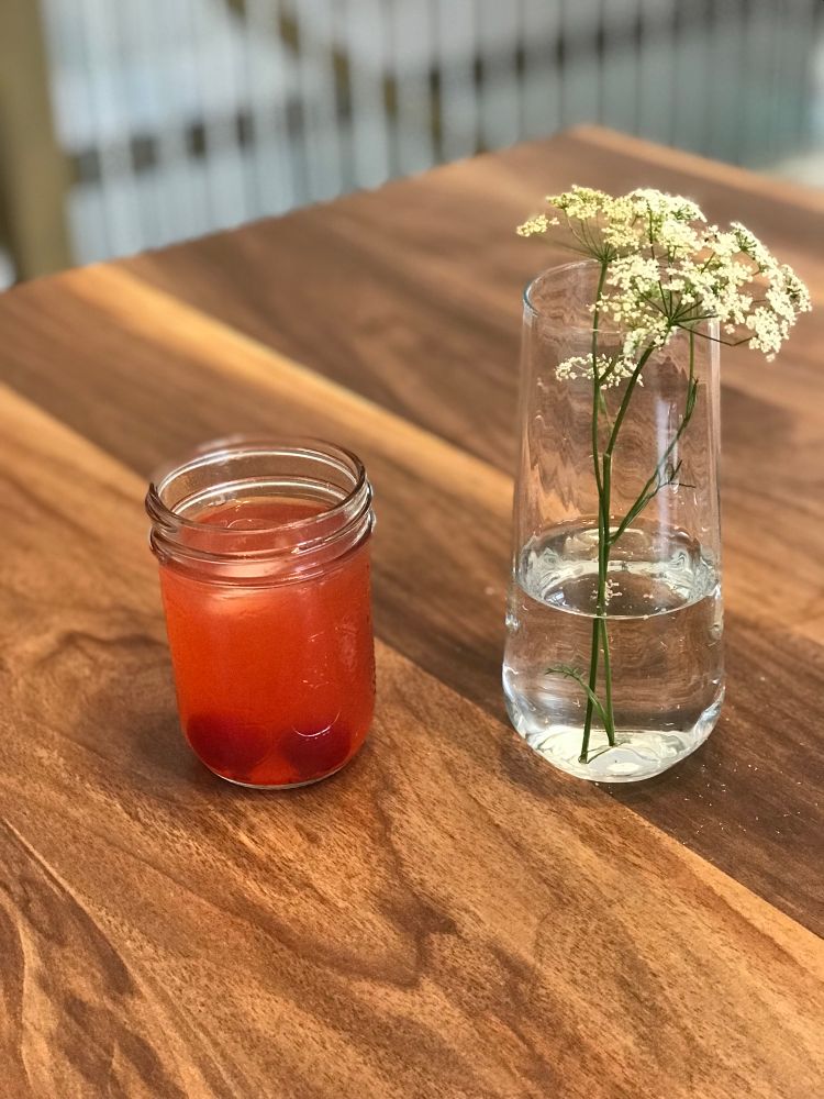 A mason jar of a cherry brandy sour with maraschino cherries beside a vase with two lacey wildflowers