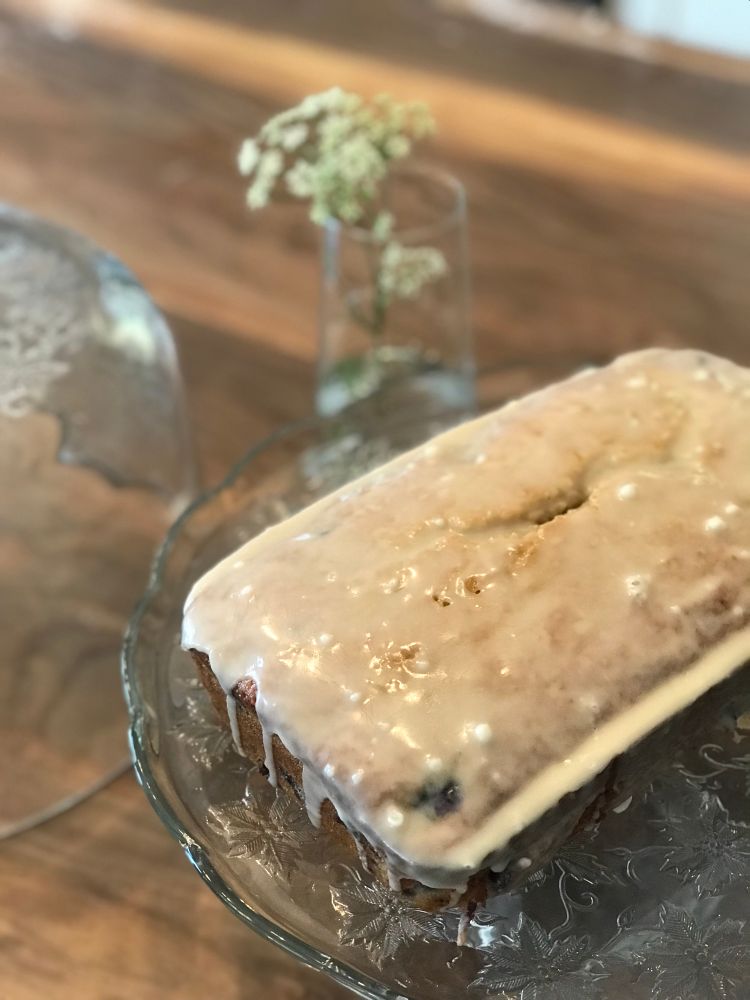 A lemon blueberry loaf dripping in lemon vanilla glaze sitting on a glass cake stand