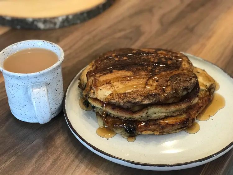 A round, ridged plate with a stack of 3 blueberry pancakes topped with syrup beside a speckled mug of tea 