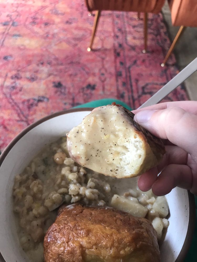 A bowl of peppery Parmesan potato and barley soup. A hand is holding up a soft pretzel bite that has been dunked in the soup.