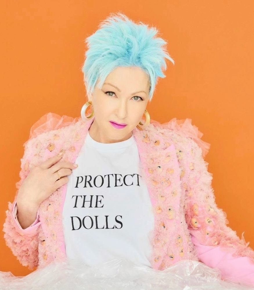 Screenshot of photo from Ms. Lauper's ig post. She's got light blue hair and fuzzy pink coat over a white t-shirt showing black letters saying PROTECT THE DOLLS 