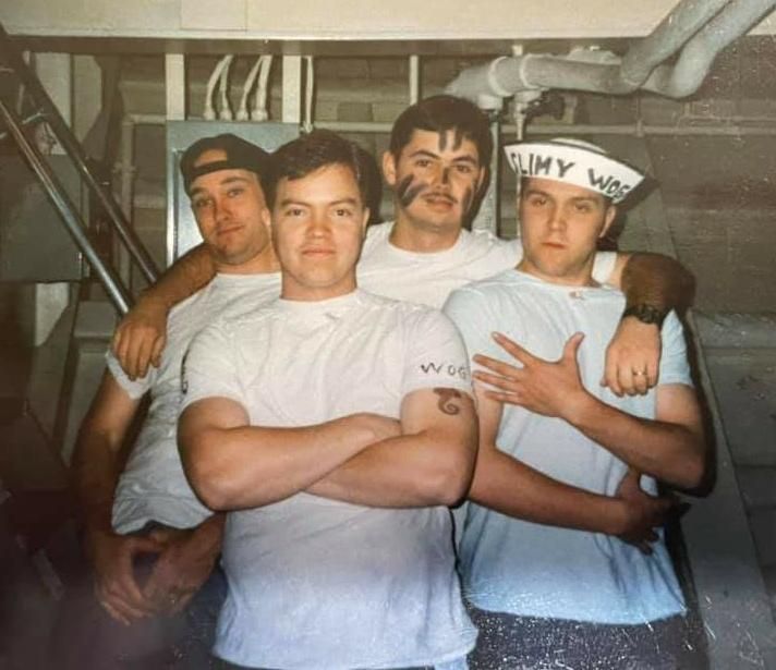 4 young white men in dungaree pants and white crew t-shirts onboard a naval ship (USS Essex). You can see random ship stuff like pipes and whatnot. 