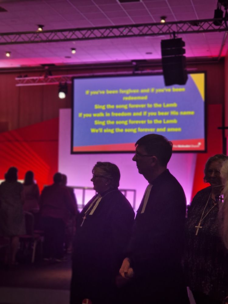 Silhouettes of the President of the Conference and Secretary of the Conference entering the hall, with lines to a hymn out of focus on a large screen behind.