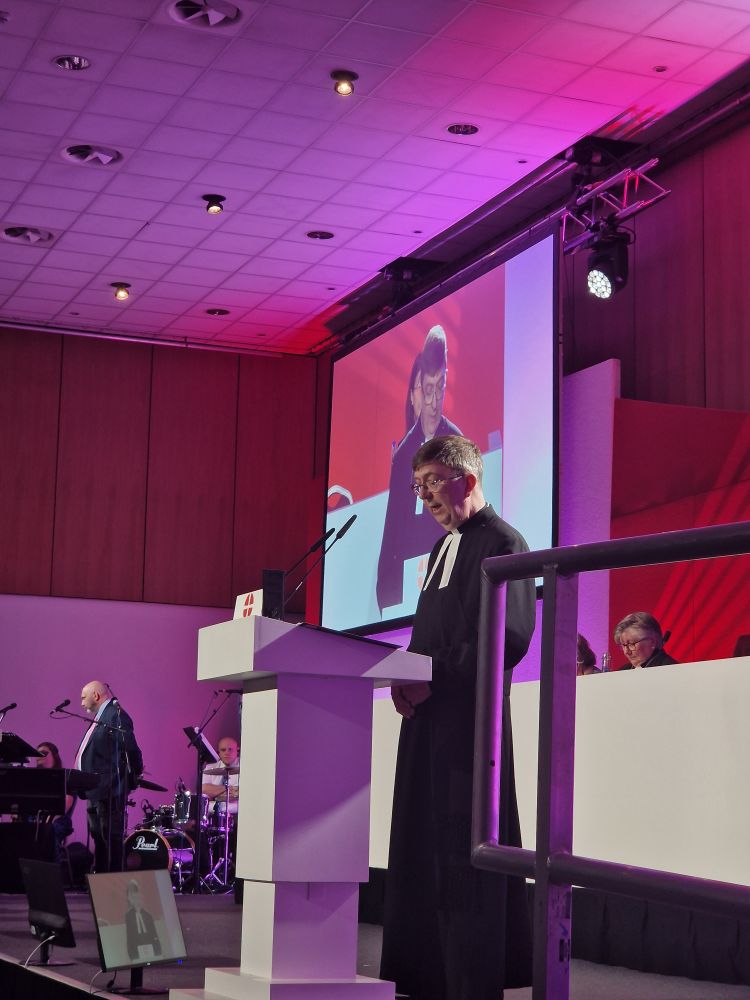 The secretary of Conference speaking at a white lectern with large screens showing a live video of the conference stage behind.