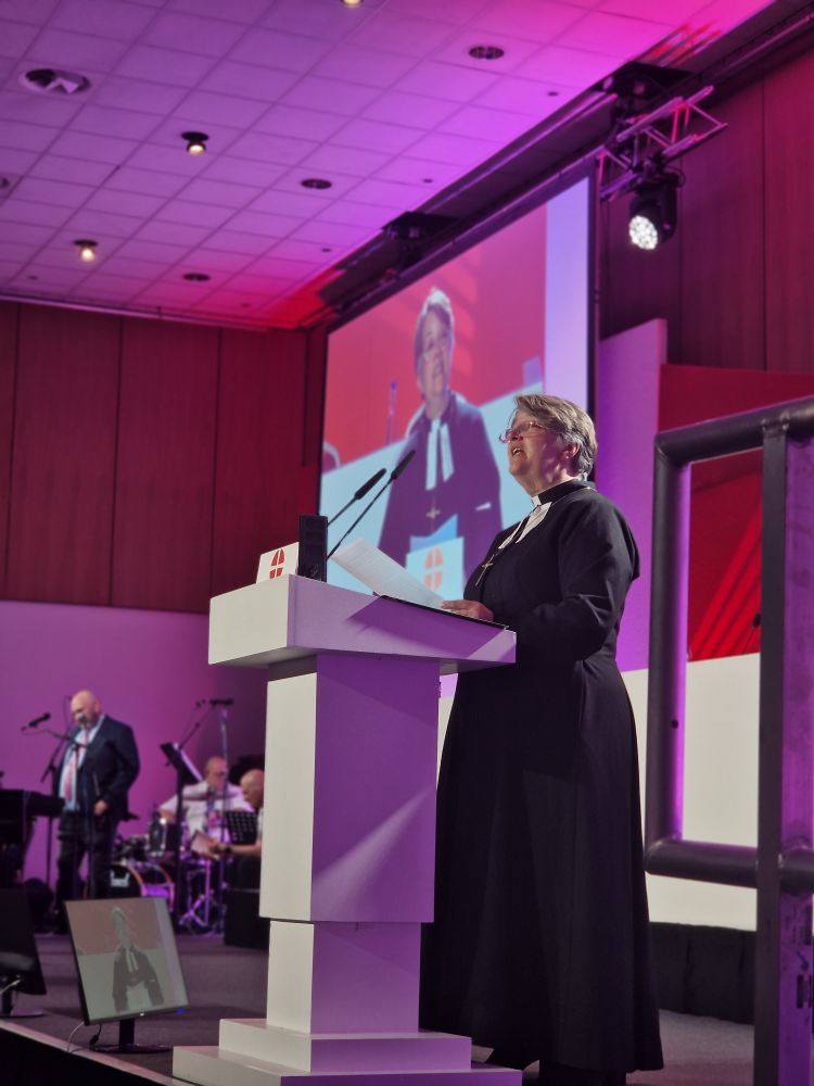 The assistant secretary of Conference speaking at a white lectern with large screens showing a live video of the conference stage behind.