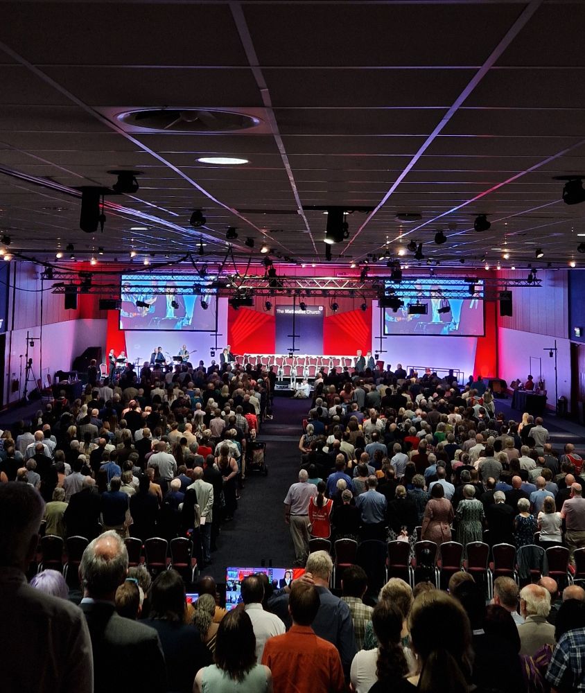 Photo of the full conference hall, looking down at rows of people facing the stage, which is illuminated by red lights and two large screens on either side. 