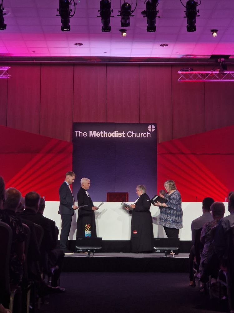 The Ex-President and Ex-Vice-President stand across from the President and Vice-President on the Conference stage, with a large Methodist logo and red backdrop  behind them.