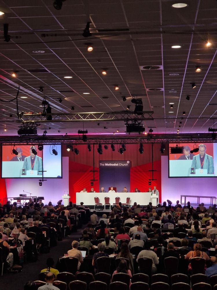 Photo of the Conference hall taken from the back of the room, showing rows of people sat facing the front, where the President and Vice President sit behind a long white desk below a large Methodist Church logo. On either side of the stage is a large screen, showing a live stream of the Conference. 