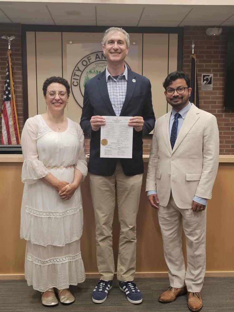 A photo of a three people in a row at Ann Arbor city hall. The two people on the ends just got married. The woman on the left is in a white silk dress, the man on the right is in a beige linen suit. The man in the center is the Ann Arbor mayor, Chris Taylor, holding the couples completed marriage certificate.