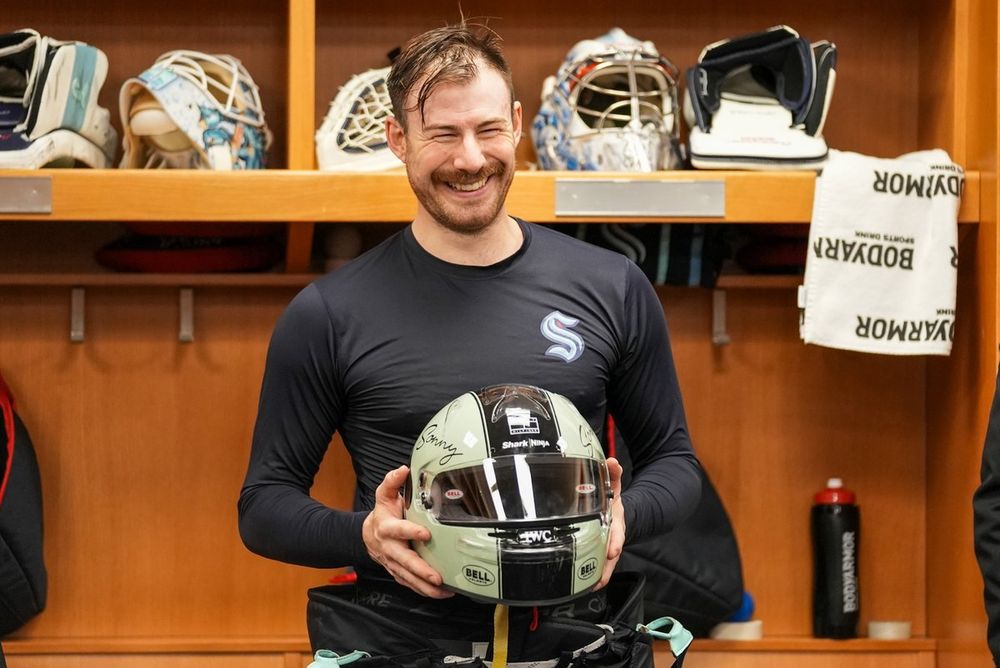 a photo of Seattle Kraken goaltender Philipp Grubauer smiling widely as he holds the team's victory helmet in his hands.