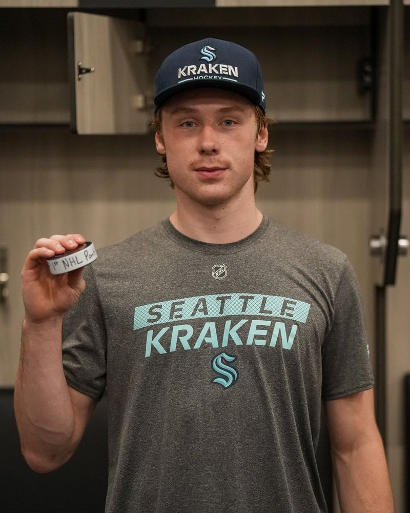 a photo of Berkly Catton in a gray Seattle Kraken t-shirt and blue cap, holding a puck that reads: 1st NHL point.