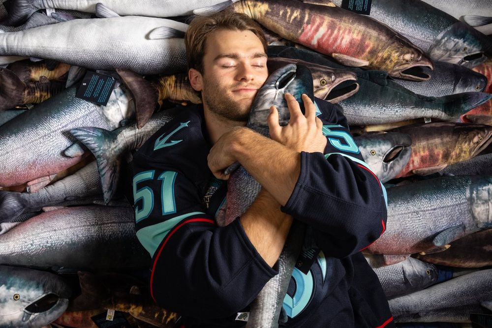 Seattle Kraken forward Shane Wright cuddles a king salmon fish plushie while lying in a pile of fish.