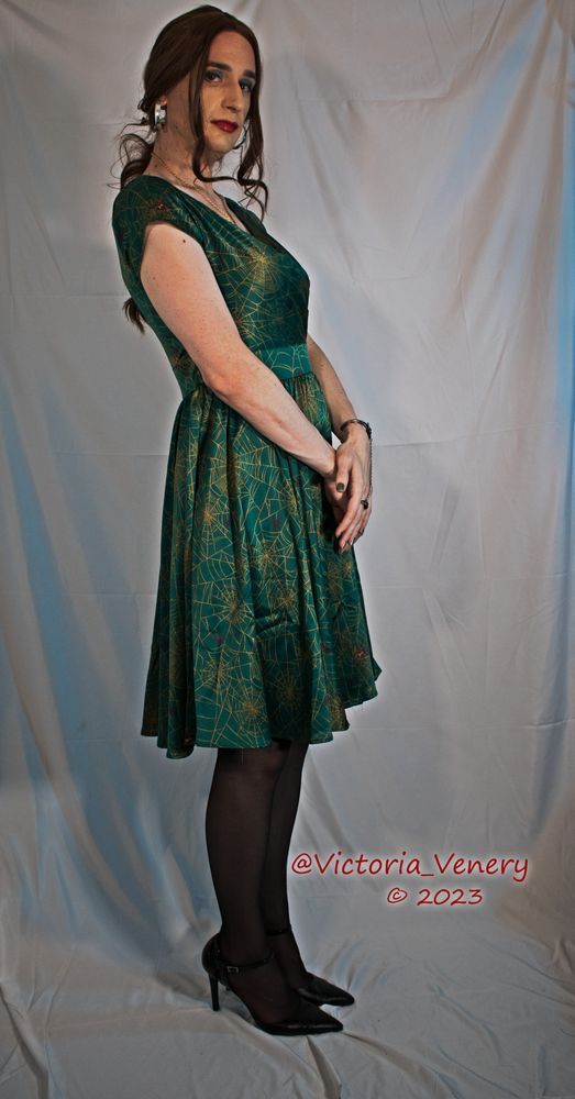 A crossdresser in a green dress with gold spiderweb pattern against a white background
