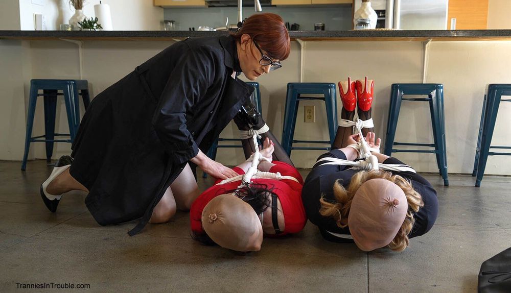 The scene takes place in a kitchen or dining area with stools lined up along a counter. Two crossdressers are lying on the floor, positioned side-by-side, both wearing stockings and outfits that suggest a costume or role-play theme. Their arms and legs appear to be tied with white rope in a deliberate, stylized fashion, and they each have nude-colored hoods covering their heads. One is wearing bright red high heels.

A third crossdresser, wearing glasses, a dark coat, stockings, and heels, is kneeling beside them, appearing to check or adjust the ropes.