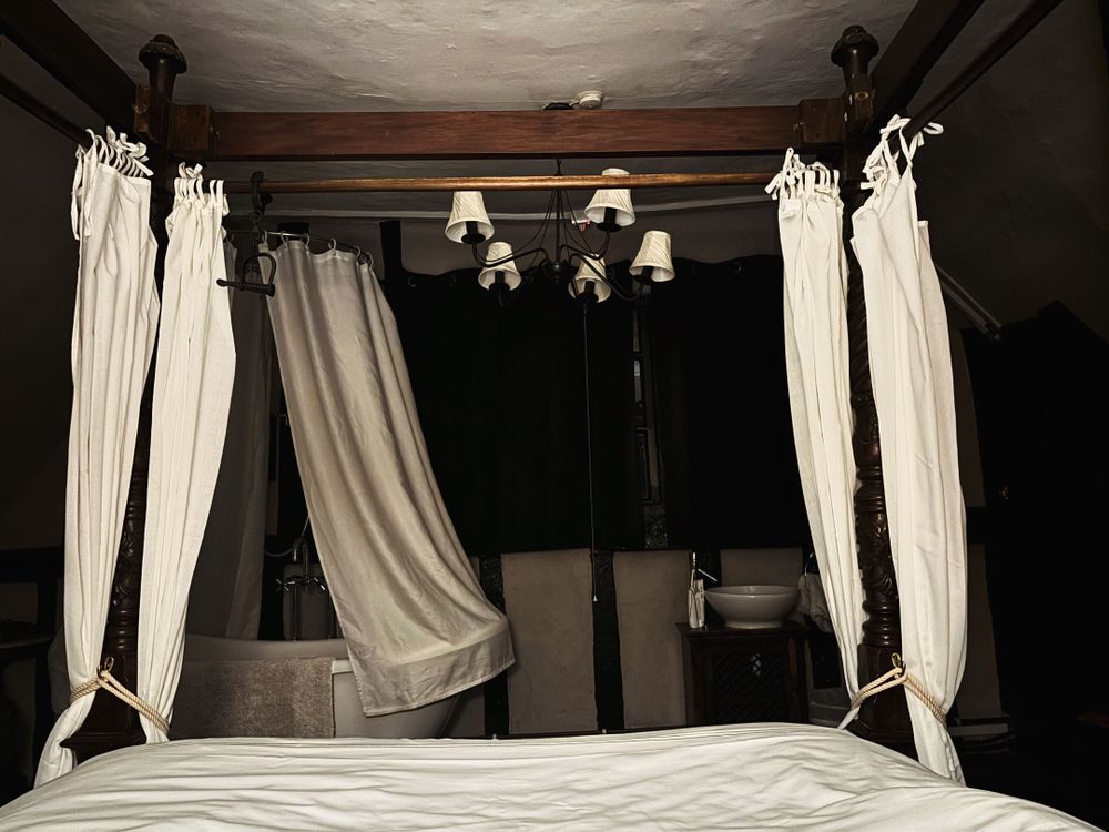 a four poster bed with white drapes. There's a bath with white drapes around it to the left and a dark window at the end of the bed. The light is eery showing an old and uneven ceiling.