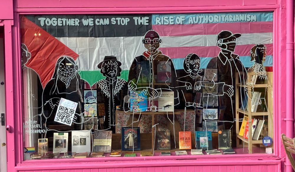 Painted windows at the Amnesty Bookshop. Shows outlines of people linked arm in arm, with text saying “Together we can stop the rise of authoritarianism” with a trans flag and a Palestinian flag behind it.