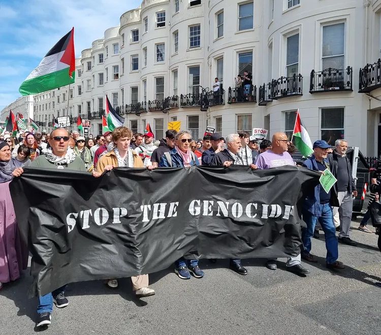 A large crowd marching behind a black banner that says STOP THE GENOCIDE. There a Palestinian flags in the crowd behind. They are marching past a street of white Georgian seafront houses in Brighton.  