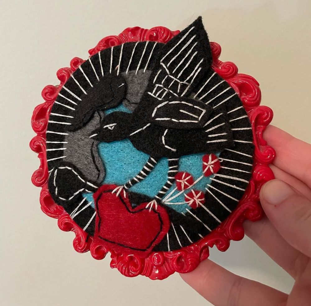 A felt and thread black bird on a teal background, holding a red heart with its feet. The piece is in a red plastic round frame with a scroll design on it.
