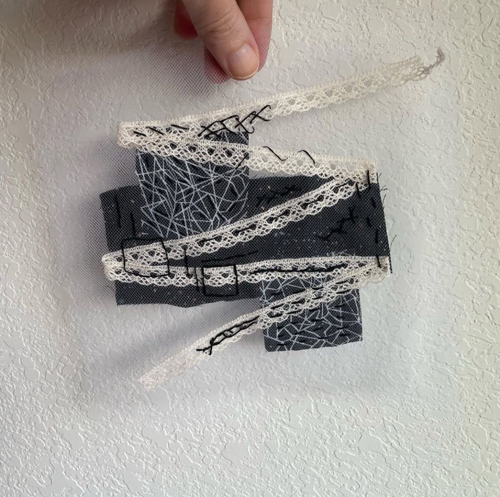 A fabric collage made of white tulle, off-white lace, and black and white patterned fabric. The collage is held up against a white wall.