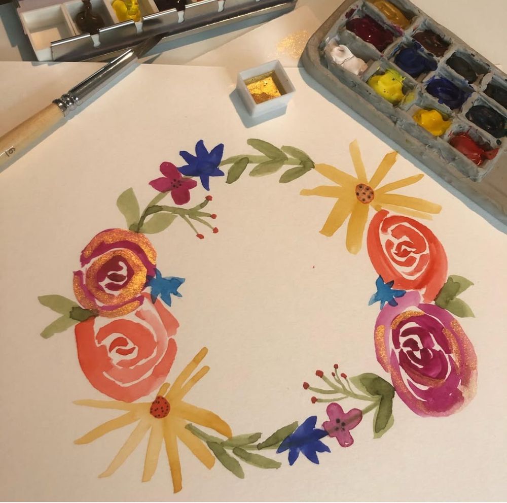 A watercolor painting of a wreath made of flowers. There are pink and orange roses, yellow daisies, and blue and pink tiny flowers with green leaves. Some of the flowers have gold accents. A tray of watercolor paints and a brush are in the background.