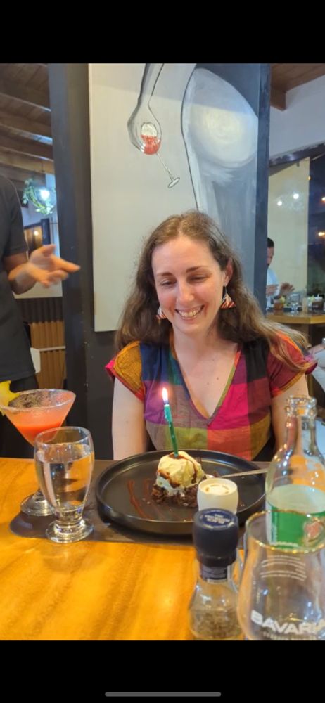 A white wkman with brown hair and a multi-colored jumpsuit looking at a candle stuck in a brownie sundae.