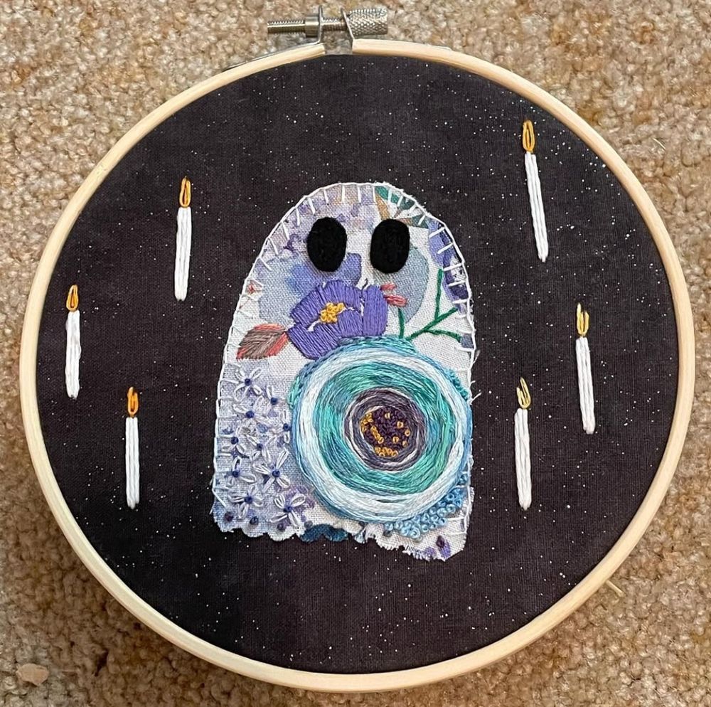 A floral ghost is stitched on sparkly black fabric and embellished with embroidered candles. The ghost is different shades of blue and purple with black felt eyes.