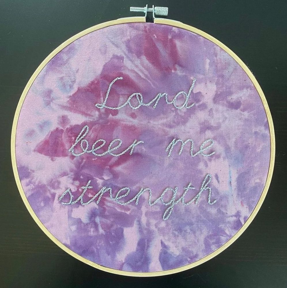 An embroidery hoop with the words “lord beer me strength” stitched in a light blue thread on ice-dyed fabric that is multiple shades of purple