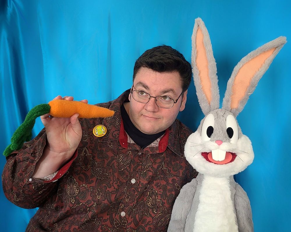 The puppet builder, in a vintage style shirt and wearing a Bugs Bunny pin, poses with the Bugs Bunny puppet built by UzzyWorks. Also holding a carrot and posing in a Groucho Marx sort of way.