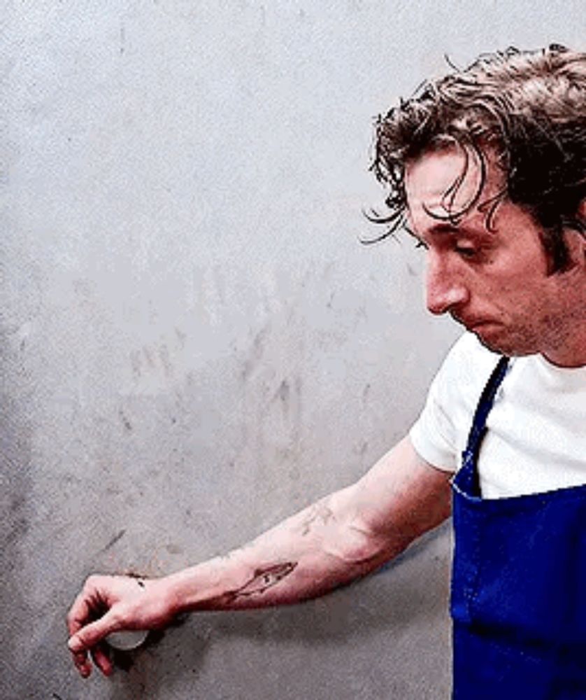 a man with a tattoo on his arm is wearing a blue apron and a white shirt .