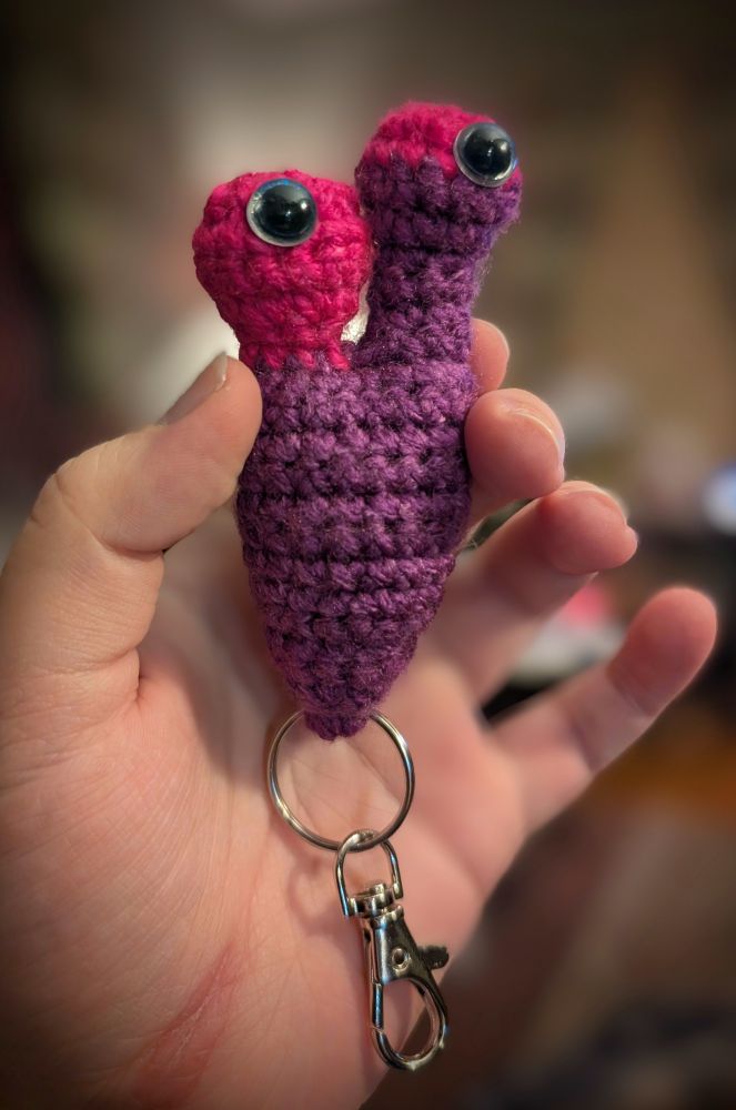A crochet slug keychain. The body is purple and one eye stock is mostly purple with pink at the top. The other eye stock is pink. It has glittery blue eyes near the top of each eye stock. A silver keyring and clip are hanging from the bottom. 