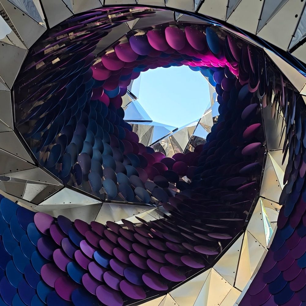 Looking up through the swirling interior of "Orbital," a mirrored art installation lined with purple and blue reflective discs, revealing a patch of blue sky at the center. The structure curves like a vortex with geometric facets catching the light.
