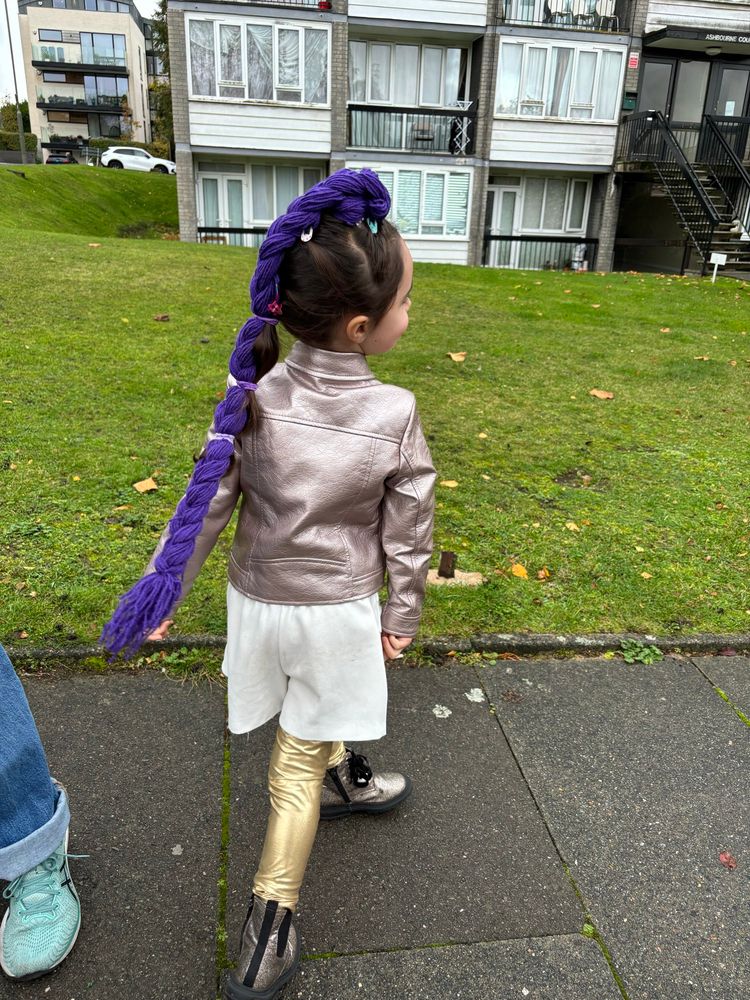 A little girl dressed in little gold boots, a lighter gold pair of trousers, white shorts, an ashen gold(ish) jacket and a long plait of woollen purple hair in her brunette hair. She’s stood in front of the grass outside a block of flats
