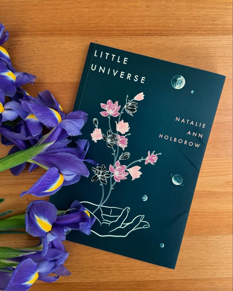 A copy of poetry collection Little Universe by Natalie Ann Holborow is placed on a a wooden table with purple irises placed on the book. 