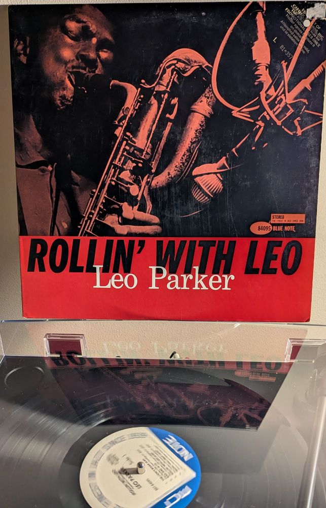 A large (top four fifths) red tinted  black and white photo of a Black man (Parker), blowing hard into his baritone sax. His cheeks are puffed out with the effort. The triple curve of the horn's neck is prominent. A large microphone is on the right. The photo is quite dark except for the light reflected off of Parker's face and his horn. The bottom fifth is red with bold black type for the title and smaller white type for the artist.