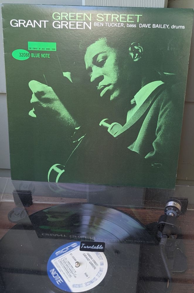 LP cover - a full frame, green tinted black and white (black and green) photo of a Black man (Green) playing his guitar, taken from near his left shoulder so we see the back of his guitar neck and just the barest bit of the white binding on the body. He's wearing a white dress shirt with the collar open, and a dark grey or black suit. There's a single light on him, illuminating his face and the knuckles of his chording hand. You can see the guitar strap coming over his shoulder and past his ear on the way to the nut. A very dark, very evocative shot.