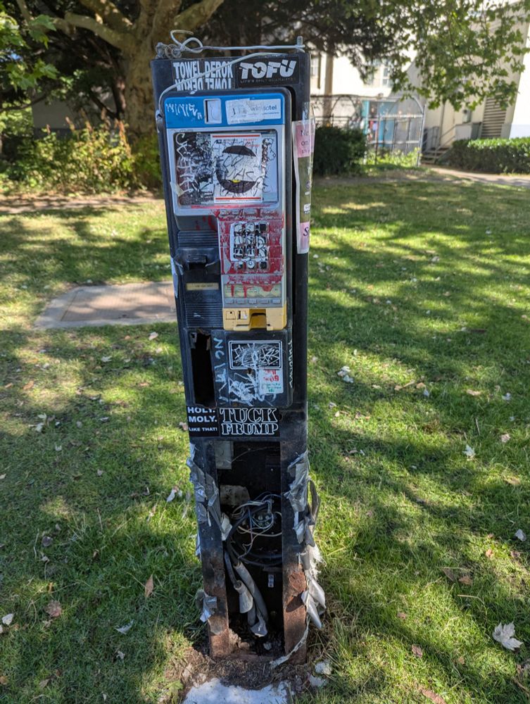 Extremely battered payphone with no handset and wiring exposed and ripped out, and covered with stickers. The best one is TUCK FRUMP.