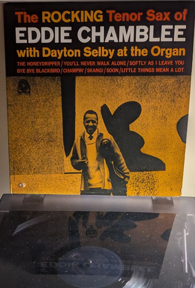 LP cover - yellow tinted black and white photo of a Black man (Chamblee) standing against a tile wall in an abstract modern art design of bold black blobs. He's holding his sax in a vinyl carrying bag, and a cigarette in his other hand. He's wearing two sweaters, a striped chunky knit cardigan over a white shawl-collar one, with a white shirt collar and a necktie underneath. He's got a big beaming smile on his face. 