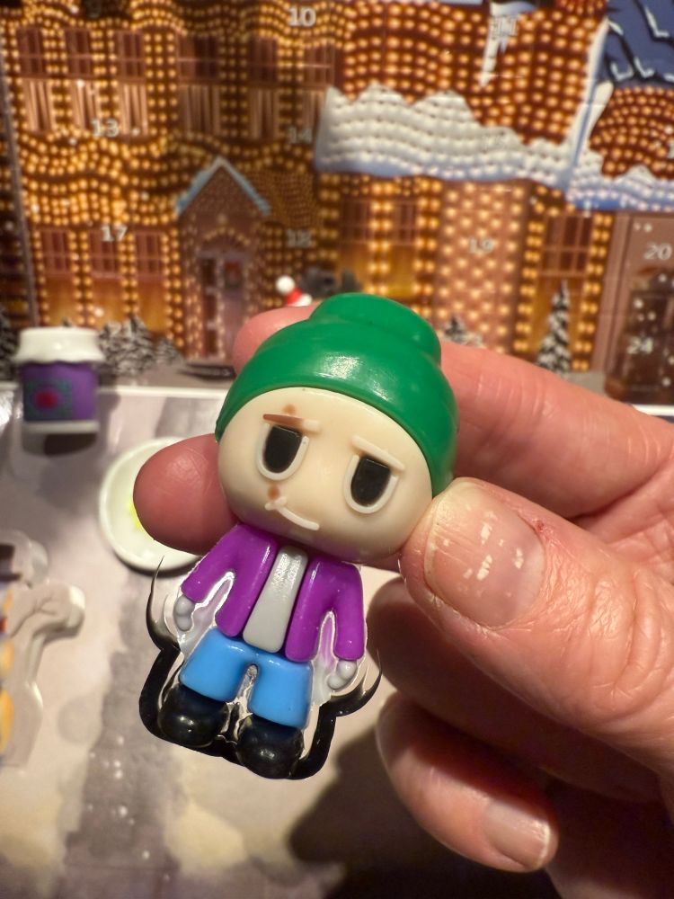Plastic figure of a white guy in a green hat? Wearing a purple cardigan over a grey shirt and blue pants. He has a brown splotch over his eye. Something appears to be connected to his lower half. A sled? 